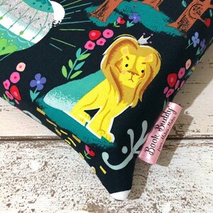 May include: A decorative pillow with a whimsical design featuring a yellow lion wearing a crown, set against a dark background with colorful flowers and foliage. The pillow has a pink tag that reads "Book Buddy from Melva Makes".