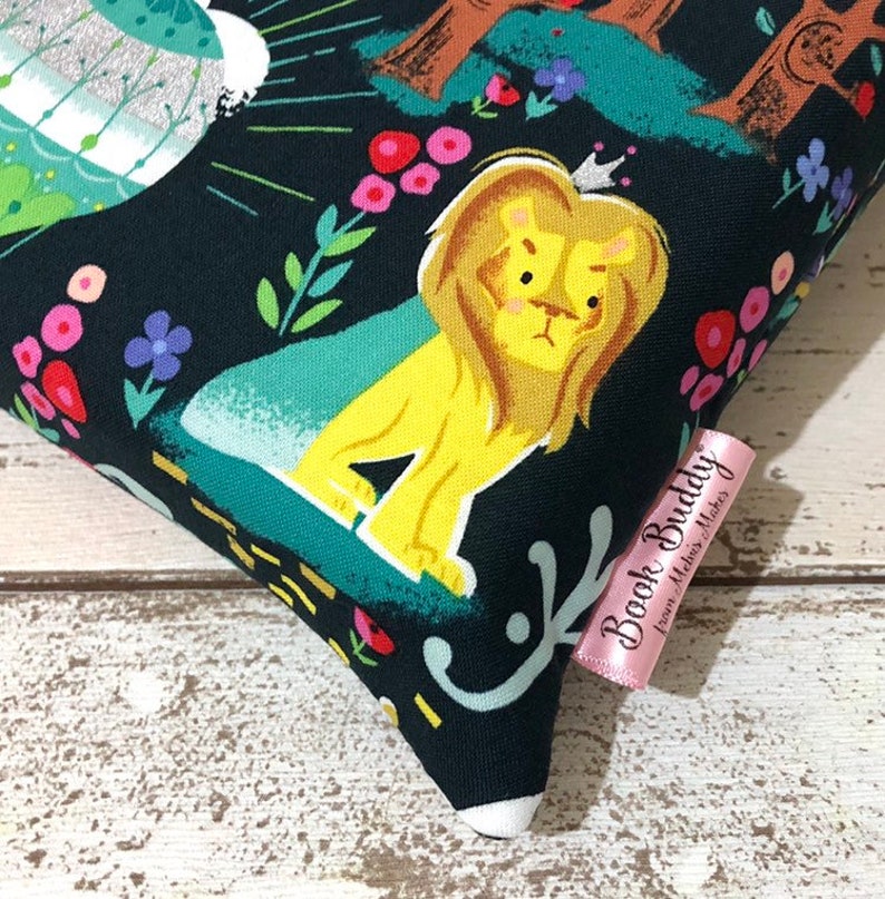 May include: A decorative pillow with a whimsical design featuring a yellow lion wearing a crown, set against a dark background with colorful flowers and foliage. The pillow has a pink tag that reads "Book Buddy from Melva Makes".