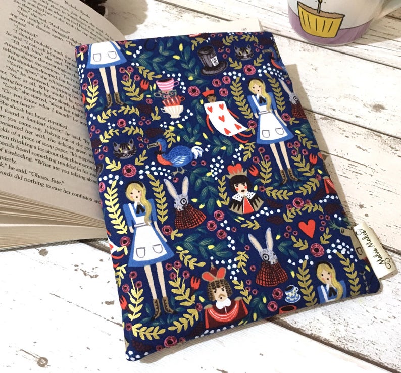 May include: A rectangular fabric book sleeve featuring a dark blue background with a repeating pattern of Alice in Wonderland characters, including Alice, the White Rabbit, the Queen of Hearts, and the Mad Hatter's hat, surrounded by foliage.