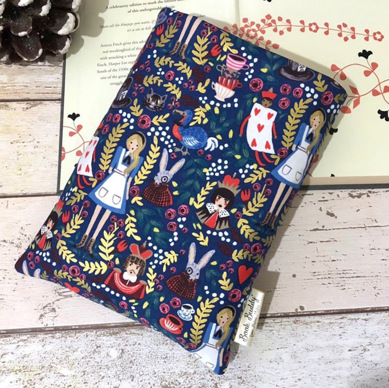 May include: A rectangular fabric book sleeve featuring a dark blue background with a repeating pattern of Alice in Wonderland characters, including Alice, the Queen of Hearts, rabbits, and teacups, with a small white tag labelled Book Buddy.