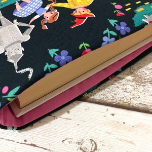 May include: A book sleeve featuring a whimsical design with characters from The Wizard of Oz. The dark blue fabric is patterned with illustrations of the Tin Man, Dorothy, and other characters, along with colorful flowers. The interior is lined with pink fabric.