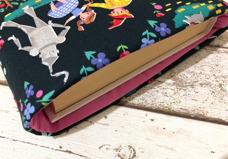 May include: A book sleeve featuring a whimsical design with characters from The Wizard of Oz. The dark blue fabric is patterned with illustrations of the Tin Man, Dorothy, and other characters, along with colorful flowers. The interior is lined with pink fabric.