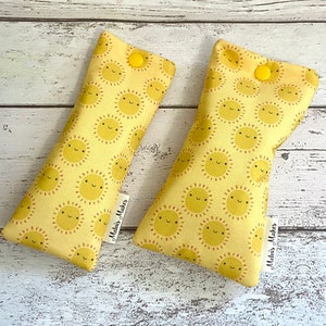 Puede incluir: Dos almohadas para ojos de tela amarilla con un patrón repetitivo de soles sonrientes. Las almohadas tienen un botón amarillo en la parte superior y una etiqueta blanca que dice "Melhuis Makes".
