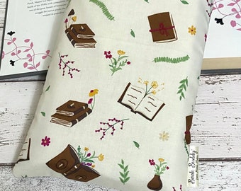May include: A white book-themed pillow with a brown book, flowers, and leaves printed on it. The pillow has a brown leather-like trim and a tag that says "Book Buddy".