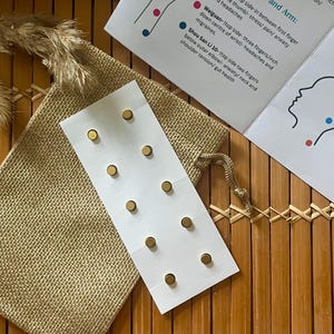 Può includere: Viene mostrata una carta bianca con dieci piccoli oggetti circolari color oro, possibilmente magneti per agopuntura. La carta è posizionata su un sacco di iuta con coulisse, accanto a un opuscolo con testo e diagrammi.