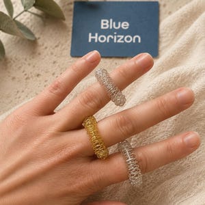 May include: Three coiled wire rings worn on fingers, featuring two silver-toned rings and one gold-toned ring. Each ring is constructed from a continuous, tightly wound metal wire coil. A blue rectangular card with the text Blue Horizon is visible.
