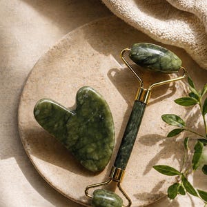 May include: A dark green jade facial roller with gold-toned metal hardware and a matching heart-shaped gua sha tool. Both items feature a smooth, polished stone surface with natural veining patterns, designed for facial massage and skincare routines.