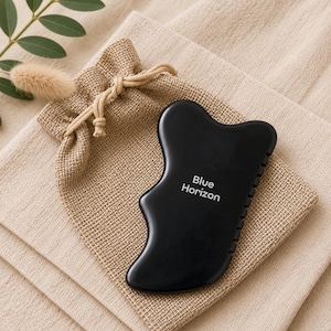 Könnte beinhalten: Ein schwarzes Gua-Sha-Werkzeug mit einer glatten, geschwungenen Form und einer gezackten Kante an einer Seite. Der Text "Blue Horizon" ist in Weiß in der Mitte des Werkzeugs aufgedruckt. Es liegt auf einem kleinen, beigen Kordelzugbeutel aus Webstoff.