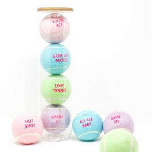 Op de afbeelding: Een doorzichtige plastic container gevuld met zes kleurrijke tennisballen. De ballen hebben verschillende zinnen erop gedrukt, waaronder "You're Ace", "Game Set Match", "Love Tennis", "Smash It", "Ace Ace Baby" en "Game On".