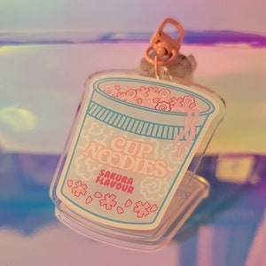 May include: A clear plastic keychain charm shaped like a cup of noodles with pink and white flowers and the text "Cup Noodles Sakura Flavour".