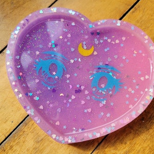 May include: A heart-shaped resin dish with a sparkly pink and purple ombre finish. The dish features a crescent moon and two blue eyes with glitter accents.
