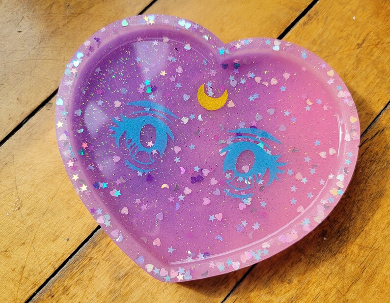 May include: A heart-shaped resin dish with a sparkly pink and purple ombre finish. The dish features a crescent moon and two blue eyes with glitter accents.