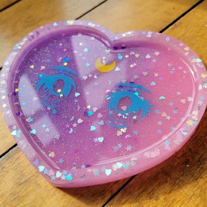 May include: A heart-shaped resin tray with a glittery purple and pink gradient. The tray features two blue eyes with eyelashes and a crescent moon.