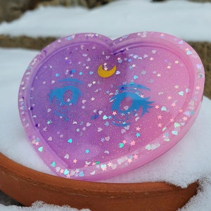May include: A heart-shaped resin tray with a purple and pink ombre finish. The tray is decorated with glitter, stars, and a crescent moon with a single eye design.