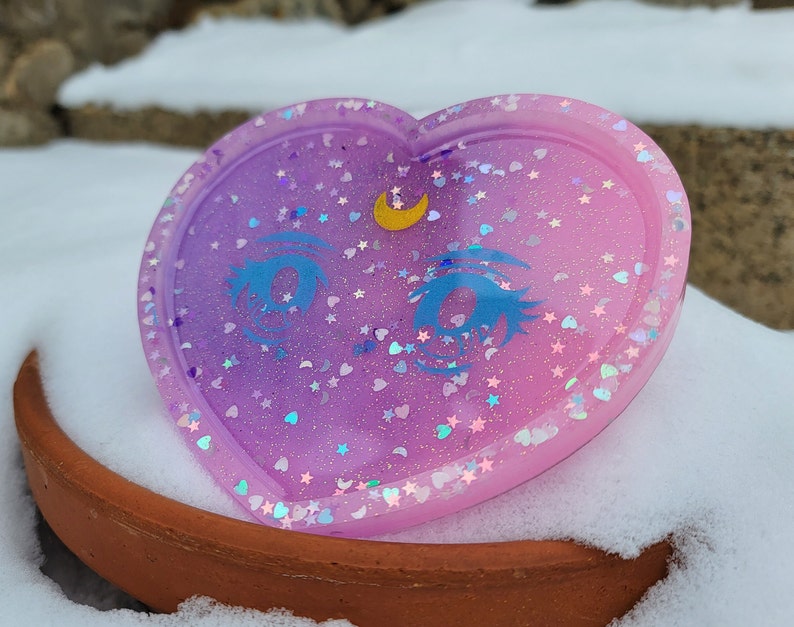 May include: A heart-shaped resin tray with a purple and pink ombre finish. The tray is decorated with glitter, stars, and a crescent moon with a single eye design.