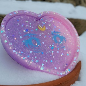May include: A heart-shaped resin coaster with a light purple and pink ombre effect. The coaster is decorated with glitter, stars, and a crescent moon with a single eye design.