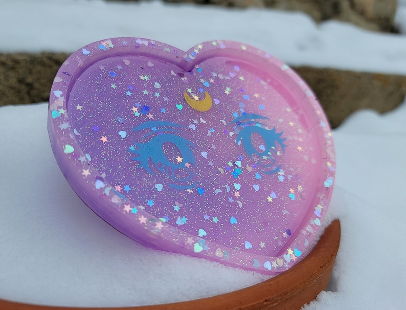 May include: A heart-shaped resin coaster with a light purple and pink ombre effect. The coaster is decorated with glitter, stars, and a crescent moon with a single eye design.