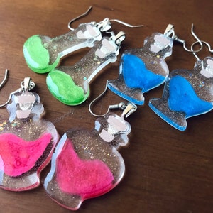 May include: Five pairs of dangle earrings featuring clear glass bottles filled with glitter and coloured liquid. The bottles are filled with pink, blue, and green liquid.