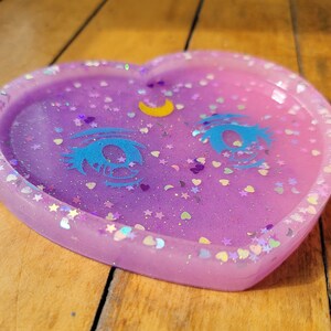 May include: A heart-shaped resin tray with a light purple and pink ombre effect. The tray is decorated with glitter, stars, hearts, and a crescent moon with a blue eye design.