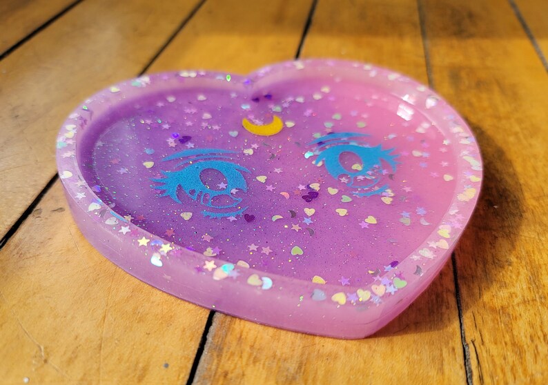 May include: A heart-shaped resin tray with a light purple and pink ombre effect. The tray is decorated with glitter, stars, hearts, and a crescent moon with a blue eye design.
