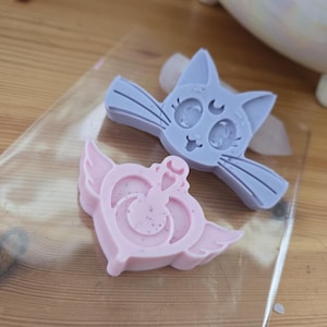 May include: Two handmade soap bars in the shape of cartoon characters. One is a pale blue cat with a crescent moon on its forehead. The other is a pink heart with wings and a crown. Both soaps are on a clear plastic sheet.