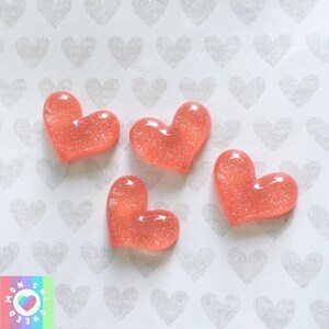 Puede incluir: Cuatro adornos de resina en forma de corazón rosa con purpurina sobre un fondo blanco con un patrón repetitivo de pequeños corazones grises.