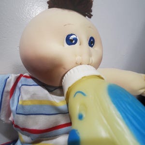 May include: A Cabbage Patch Kid doll with brown yarn hair wearing a blue and white striped shirt is drinking from a yellow plastic bottle with a blue dog design.