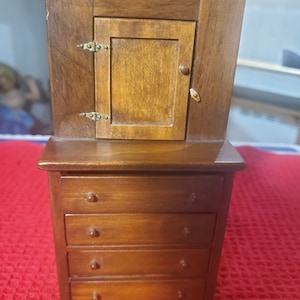 May include: A miniature wooden dresser with four drawers and a cabinet with a single door on top. The dresser and cabinet are both a dark brown colour.