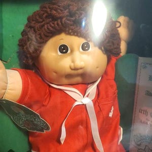 May include: A Cabbage Patch Kid doll with brown curly hair, wearing a red shirt with a white sailor collar and a white ribbon. The doll has a rosy complexion and large brown eyes.