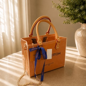 May include: A transparent orange tote bag with tan leather handles, gold-tone hardware, and a gold chain shoulder strap. A blue ribbon with the text LOUIS VUITTON is tied to the handle. The bag features the text LOUIS VUITTON MAISON FONDEE EN 1854 PARIS.