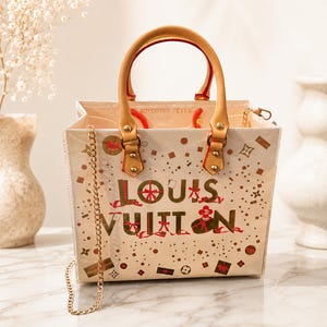 May include: A cream-colored tote bag featuring the text LOUIS VUITTON in red and gold, decorated with small gift box and star graphics. The bag has tan leather handles, gold-tone hardware, a detachable gold chain strap, and red interior accents.