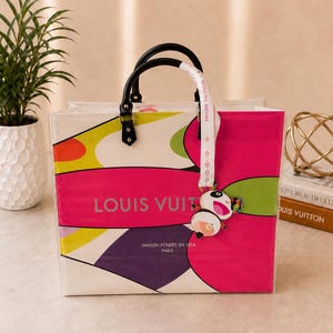 May include: A rectangular tote bag featuring a vibrant abstract pattern in hot pink, lime green, and purple. The bag displays the text LOUIS VUITTON and MAISON FONDÉE EN 1854 PARIS. It has black handles and a white ribbon charm with a panda graphic.