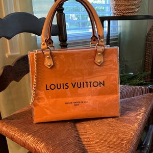May include: A square-shaped orange handbag with a clear plastic outer layer. The bag features tan leather handles with gold-tone hardware, a gold chain strap, and the black text LOUIS VUITTON and MAISON FONDEE EN 1854 PARIS printed on the front.