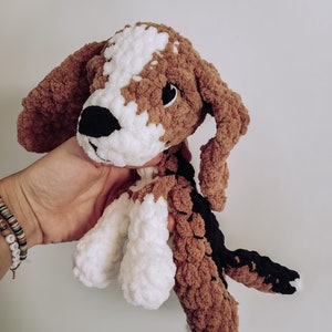 May include: A crocheted plush toy dog with brown, white, and black fur. The dog has a white face with black eyes and a black nose.