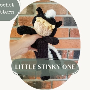 May include: A crocheted skunk amigurumi toy with black and white fur, a black nose, and a yellow face. The toy is being held by a hand in front of a brick wall. The text "LITTLE STINKY ONE" is written below the toy.