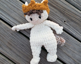 May include: A white crocheted stuffed animal wearing a brown and orange crown. The animal has a long tail and is lying on a wooden surface.