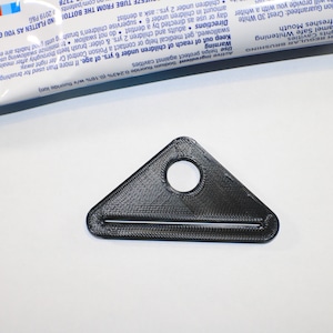 Puede incluir: Un exprimidor de tubos de pasta de dientes triangular negro con un agujero circular y una ranura para el tubo. El exprimidor está hecho de plástico y está diseñado para aplanar el tubo.