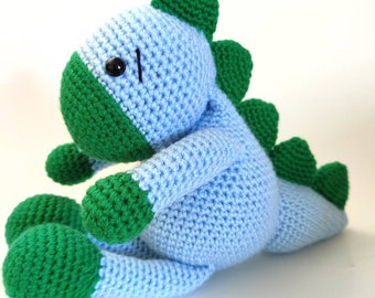 May include: A crocheted blue and green dinosaur toy. The dinosaur is sitting on a white surface.