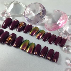 Puede incluir: Un conjunto de u&ntilde;as postizas de ata&uacute;d de color burdeos con detalles iridiscentes verdes y dorados. Algunas u&ntilde;as tienen peque&ntilde;os diamantes de imitaci&oacute;n transparentes.