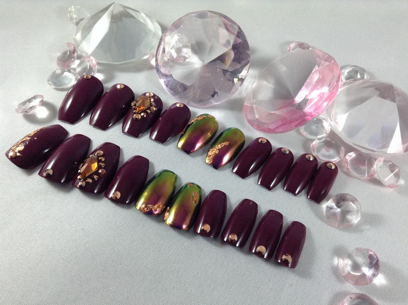 Puede incluir: Un conjunto de u&ntilde;as postizas de ata&uacute;d de color burdeos con detalles iridiscentes verdes y dorados. Algunas u&ntilde;as tienen peque&ntilde;os diamantes de imitaci&oacute;n transparentes.