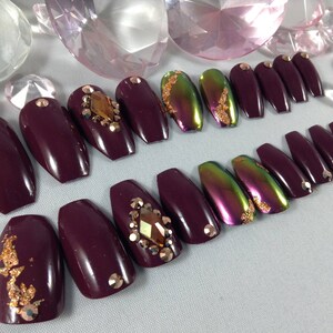 Puede incluir: Conjunto de u&ntilde;as postizas de ata&uacute;d color burdeos con detalles dorados y pedrer&iacute;a.