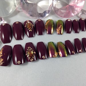 Puede incluir: Conjunto de 10 u&ntilde;as postizas de ata&uacute;d color burdeos con detalles dorados y pedrer&iacute;a.