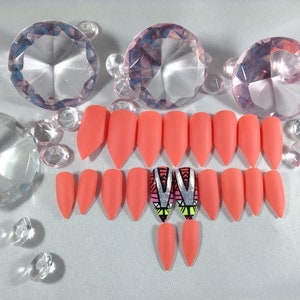 Puede incluir: Un conjunto de 12 uñas postizas de color coral rosa en forma de stiletto con un diseño geométrico en dos de las uñas.