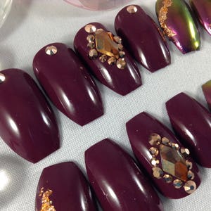 Puede incluir: U&ntilde;as postizas de ata&uacute;d color burdeos con detalles dorados e iridiscentes. Algunas u&ntilde;as tienen pedrer&iacute;a y otras tienen pan de oro.