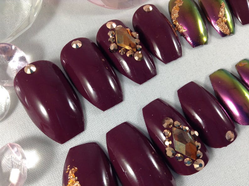 Puede incluir: U&ntilde;as postizas de ata&uacute;d color burdeos con detalles dorados e iridiscentes. Algunas u&ntilde;as tienen pedrer&iacute;a y otras tienen pan de oro.