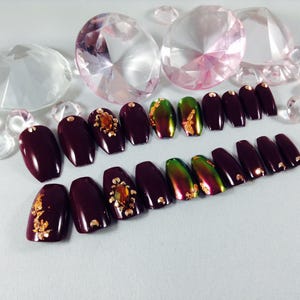 Puede incluir: Un conjunto de u&ntilde;as postizas de ata&uacute;d de color burdeos con detalles dorados. Algunas u&ntilde;as tienen un acabado verde iridiscente y otras tienen detalles de purpurina dorada.