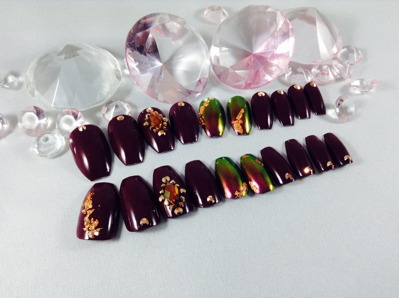 Puede incluir: Un conjunto de u&ntilde;as postizas de ata&uacute;d de color burdeos con detalles dorados. Algunas u&ntilde;as tienen un acabado verde iridiscente y otras tienen detalles de purpurina dorada.