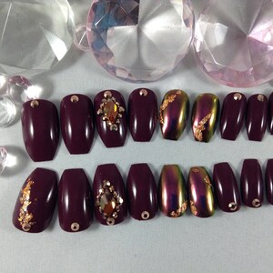 Puede incluir: Conjunto de 10 u&ntilde;as postizas de ata&uacute;d color burdeos con detalles dorados y pedrer&iacute;a. Algunas u&ntilde;as tienen un acabado hologr&aacute;fico.
