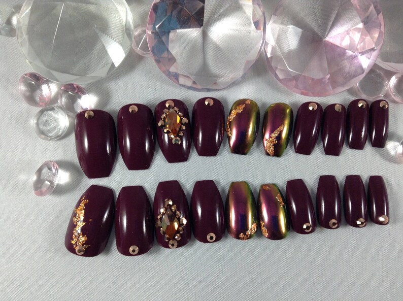Puede incluir: Conjunto de 10 u&ntilde;as postizas de ata&uacute;d color burdeos con detalles dorados y pedrer&iacute;a. Algunas u&ntilde;as tienen un acabado hologr&aacute;fico.