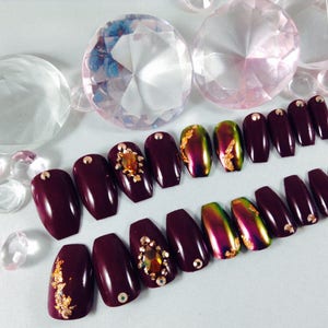 Puede incluir: U&ntilde;as postizas de ata&uacute;d color burdeos con detalles dorados y pedrer&iacute;a. Algunas u&ntilde;as tienen un acabado hologr&aacute;fico.